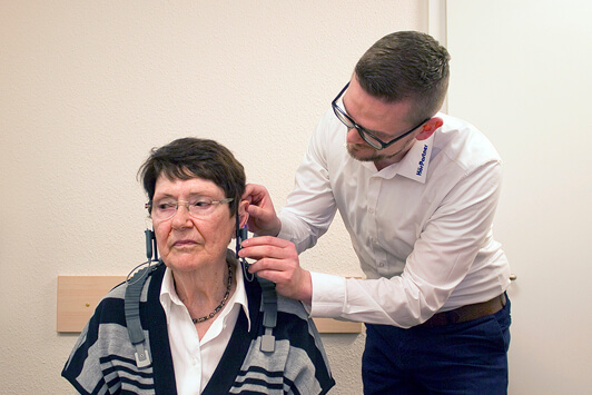 Ein Hörakustiker passt einer älteren Frau ein Hörgerät zur Hörtestberatung an.